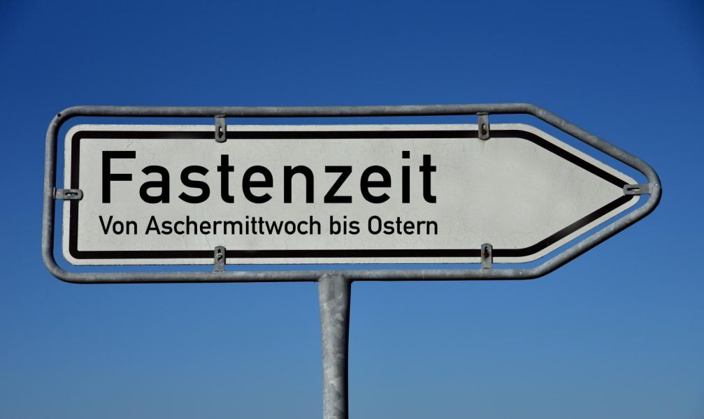 Weißer Wegweiser mit Rahmen und Fuß aus Metall vor blauem Himmel. Aufschrift in schwarzer Schrift: Fastenzeit. Von Aschermittwoch bis Ostern.