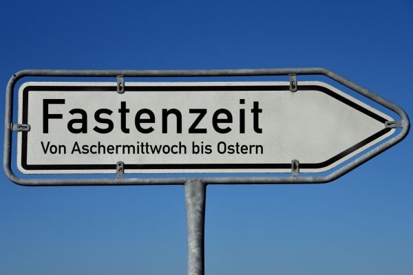 Weißer Wegweiser mit Rahmen und Fuß aus Metall vor blauem Himmel. Aufschrift in schwarzer Schrift: Fastenzeit. Von Aschermittwoch bis Ostern.
